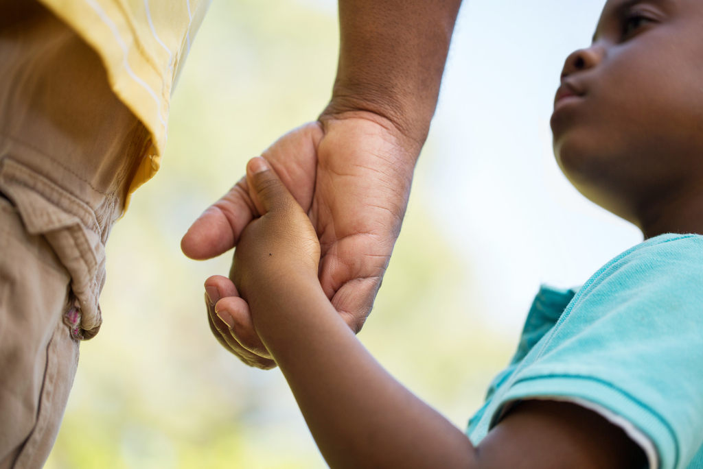 A father and son hold hands and we focus on the closeup of their hands. The boy might be 5, and he looks up slightly at his father. You can only see dad's shorts and a bit of his shirt. It's a comforting image.