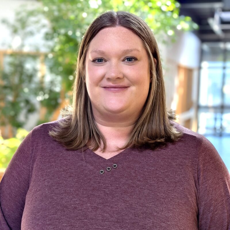 Headshot of a woman inside an atrium with tall plants. She's smiling slightly and looks calm.